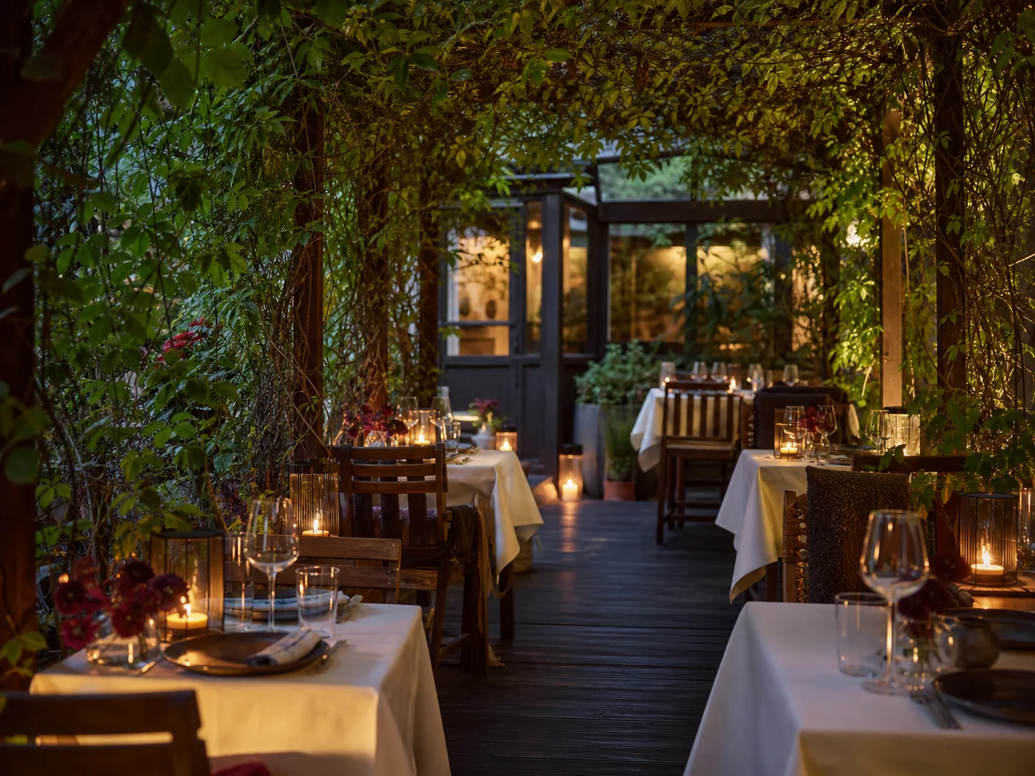 Patio restauracji Culinary Patio wieczorem, z drewnianymi stołami, lampionami, bujną zielenią i delikatnymi światłami girlandowymi.